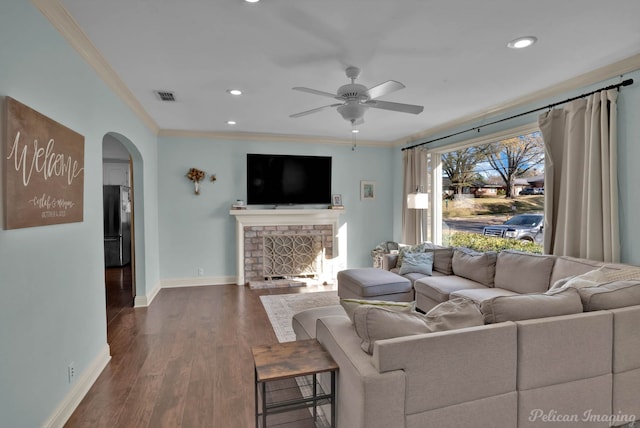 living room featuring a brick fireplace, dark wood-type flooring, crown molding, and ceiling fan