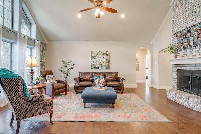 living room with wood-type flooring, ceiling fan, high vaulted ceiling, a brick fireplace, and crown molding