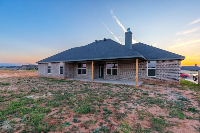 back of house at dusk featuring a shingled roof, a patio area, brick siding, and a chimney