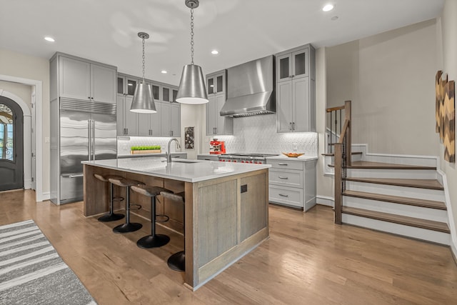 kitchen featuring decorative light fixtures, wall chimney range hood, sink, an island with sink, and stainless steel appliances