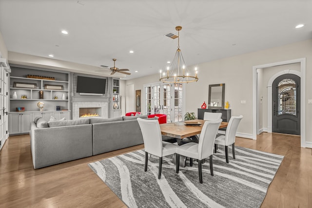 dining area with ceiling fan with notable chandelier, french doors, light hardwood / wood-style floors, a large fireplace, and built in features