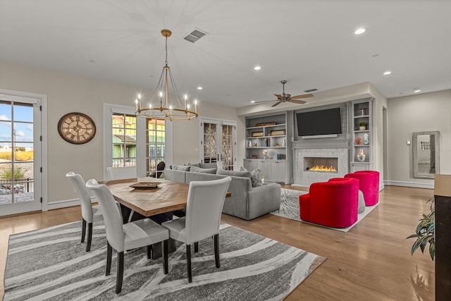 dining area featuring light wood-type flooring, ceiling fan with notable chandelier, a tiled fireplace, and built in shelves