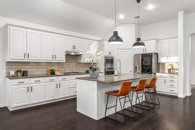 kitchen featuring white cabinets, appliances with stainless steel finishes, dark stone countertops, an island with sink, and hanging light fixtures