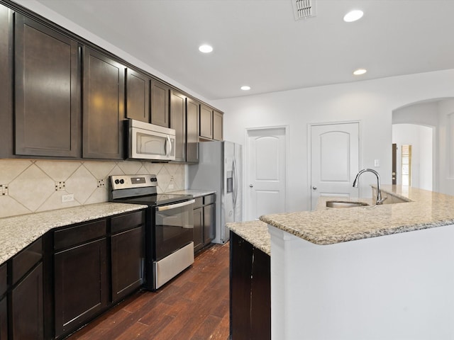 kitchen with appliances with stainless steel finishes, dark wood-type flooring, sink, a kitchen island with sink, and light stone counters