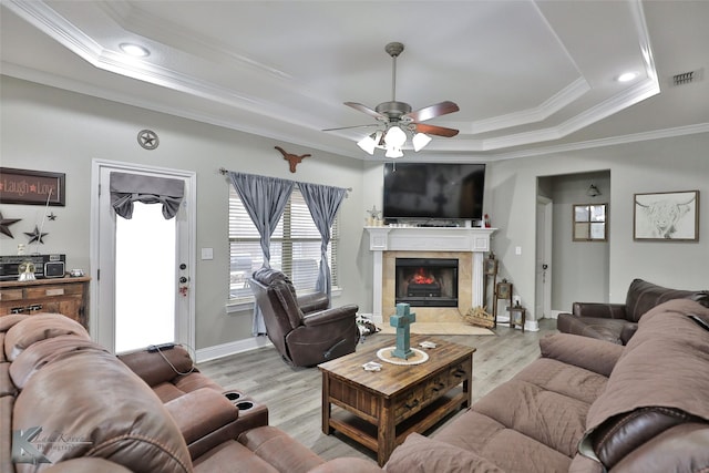 living room featuring ceiling fan, a tiled fireplace, a raised ceiling, crown molding, and light hardwood / wood-style flooring