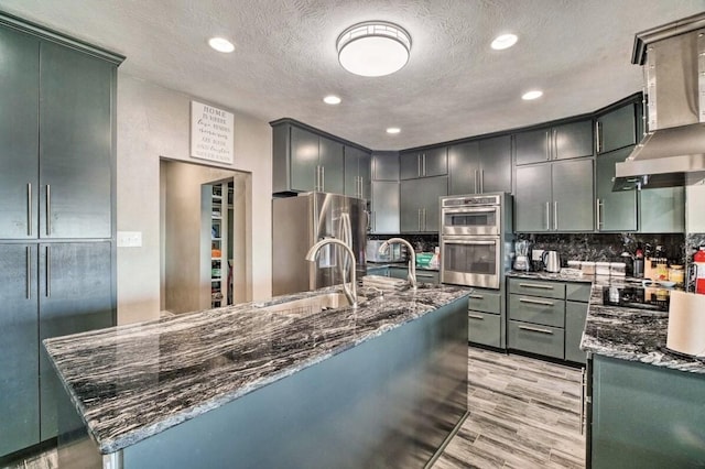 kitchen with stainless steel appliances, wall chimney exhaust hood, dark stone counters, and sink