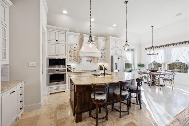 kitchen featuring hanging light fixtures, appliances with stainless steel finishes, a kitchen island with sink, a sink, and light stone countertops