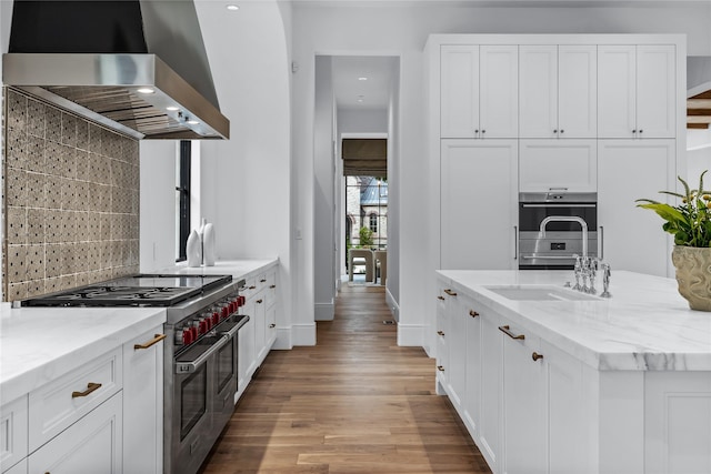 kitchen with light stone countertops, wall chimney exhaust hood, stainless steel appliances, and white cabinetry