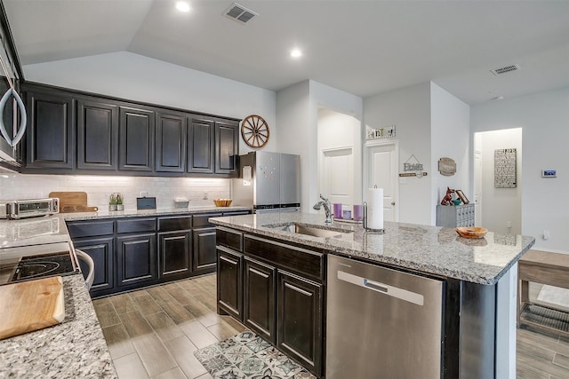 kitchen featuring stainless steel appliances, a kitchen island with sink, vaulted ceiling, light stone counters, and sink