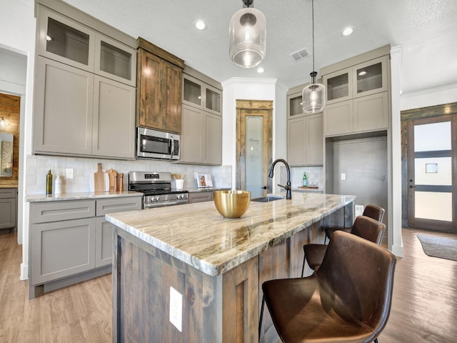 kitchen with appliances with stainless steel finishes, decorative backsplash, decorative light fixtures, an island with sink, and light stone counters