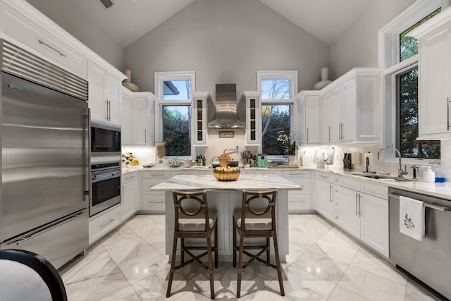 kitchen featuring white cabinetry, a center island, wall chimney range hood, built in appliances, and sink