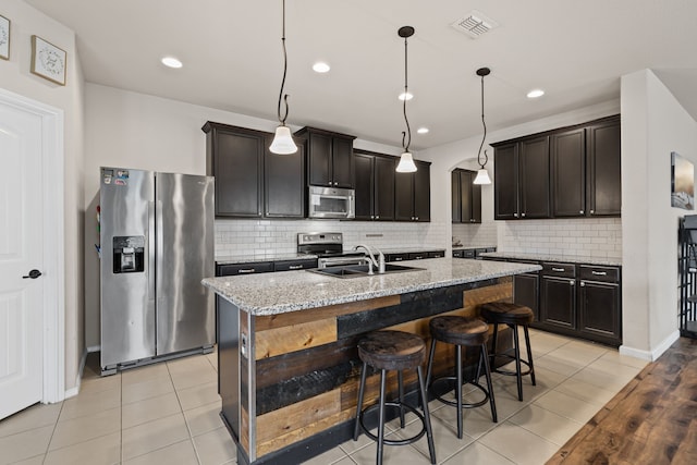 kitchen featuring hanging light fixtures, a kitchen island with sink, stainless steel appliances, light stone countertops, and dark brown cabinets