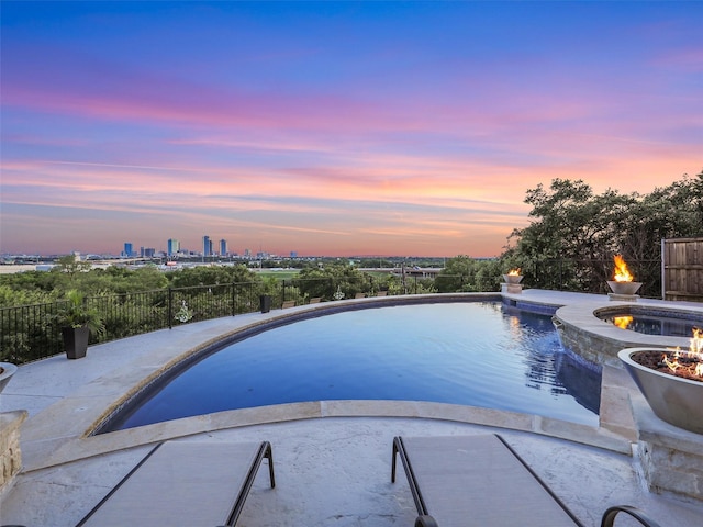 pool at dusk with a patio area, a city view, an outdoor fire pit, and fence