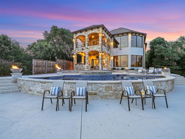 back of house at dusk with stone siding, a balcony, a patio, and fence