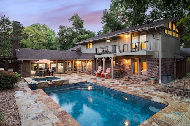 back house at dusk featuring a pool with hot tub, a balcony, and a patio