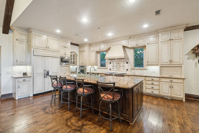 kitchen featuring a center island with sink, built in appliances, a kitchen breakfast bar, custom range hood, and cream cabinets