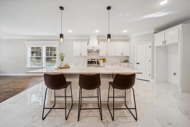 kitchen featuring light stone countertops, premium range hood, white cabinets, decorative light fixtures, and an island with sink