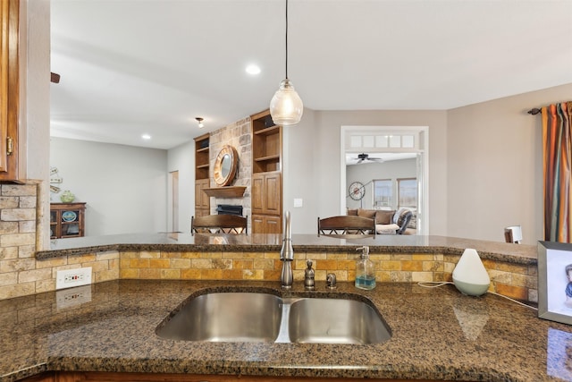 kitchen featuring a stone fireplace, decorative light fixtures, built in features, dark stone counters, and sink