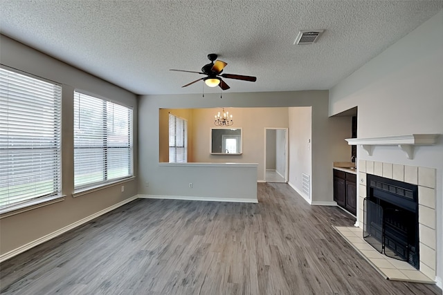 unfurnished living room with a tiled fireplace, ceiling fan with notable chandelier, a textured ceiling, and light hardwood / wood-style floors