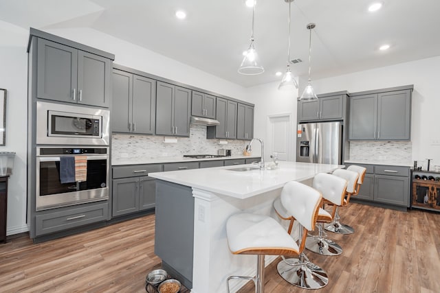 kitchen with sink, gray cabinetry, a kitchen island with sink, stainless steel appliances, and a kitchen bar