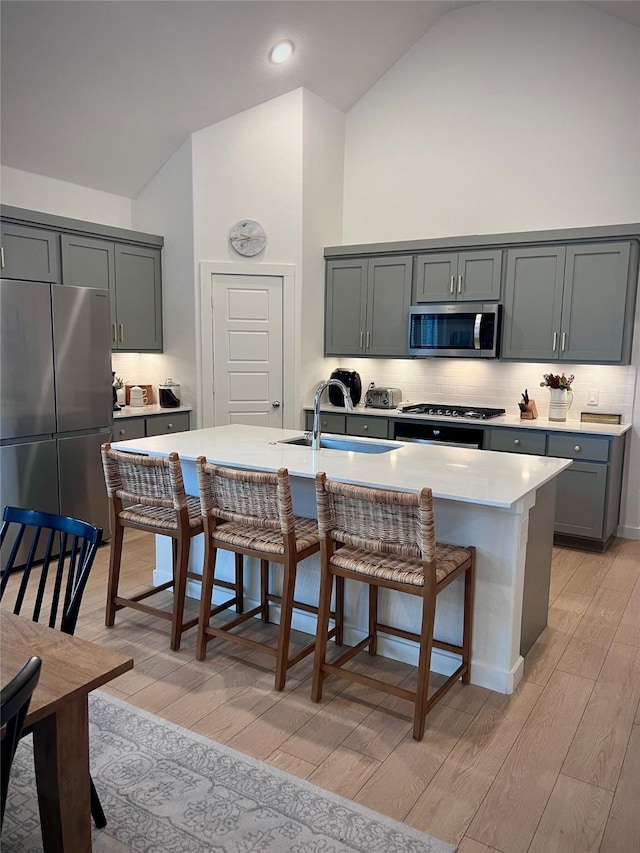 kitchen featuring sink, light hardwood / wood-style flooring, appliances with stainless steel finishes, high vaulted ceiling, and a center island with sink