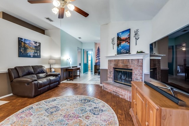 living room featuring ceiling fan, wood finished floors, visible vents, baseboards, and a brick fireplace