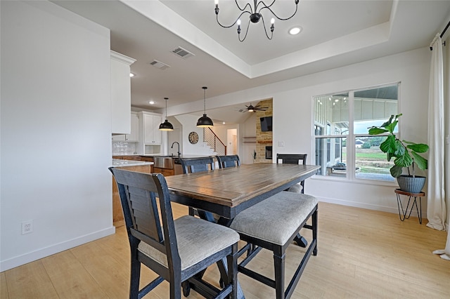 dining area featuring light wood-type flooring, a large fireplace, ceiling fan with notable chandelier, and a tray ceiling