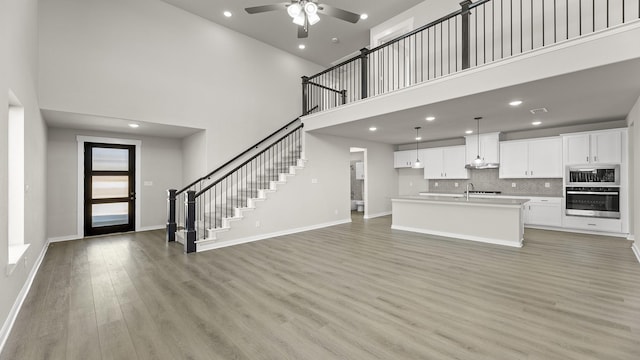 unfurnished living room featuring a high ceiling, a ceiling fan, baseboards, stairs, and light wood-style floors