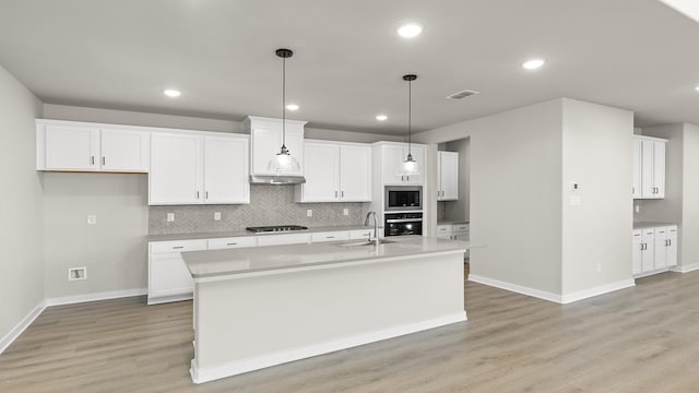 kitchen featuring wall oven, a kitchen island with sink, white cabinets, light countertops, and stainless steel microwave