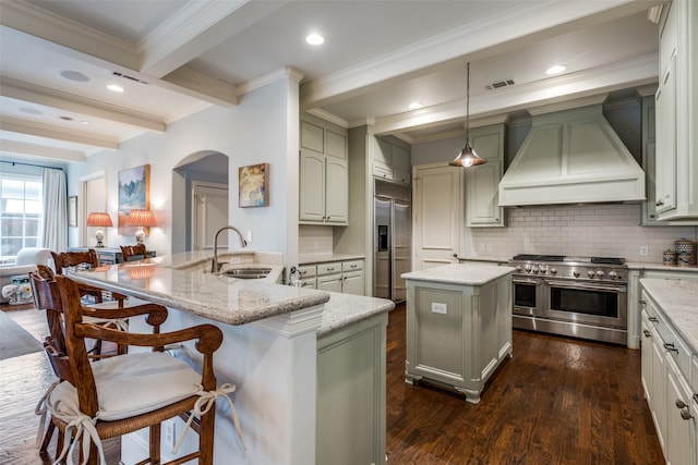 kitchen featuring custom exhaust hood, beam ceiling, sink, premium appliances, and an island with sink