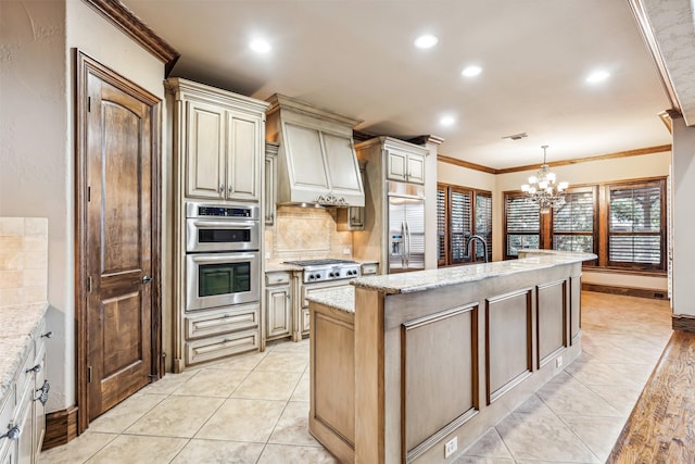 kitchen featuring decorative light fixtures, a center island with sink, stainless steel appliances, cream cabinetry, and light stone counters