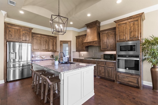 kitchen with stainless steel appliances, custom range hood, a kitchen island with sink, light stone countertops, and sink