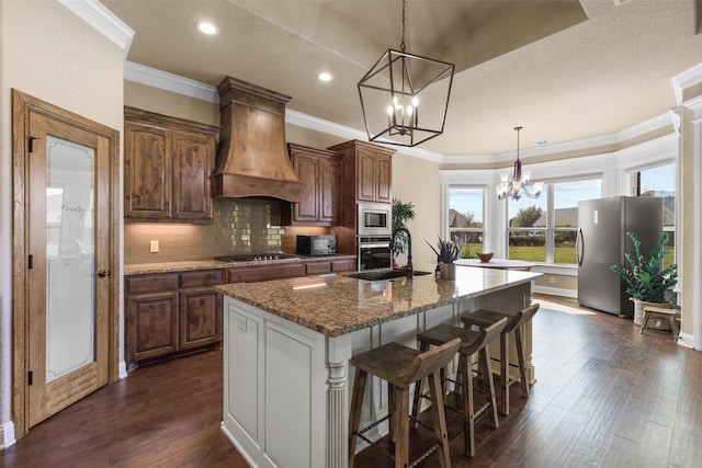 kitchen featuring pendant lighting, appliances with stainless steel finishes, an inviting chandelier, an island with sink, and premium range hood