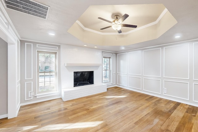 unfurnished living room featuring a fireplace, a raised ceiling, light hardwood / wood-style flooring, ornamental molding, and ceiling fan