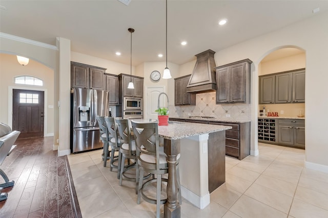 kitchen with sink, appliances with stainless steel finishes, a kitchen island with sink, dark brown cabinetry, and custom exhaust hood