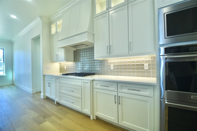 kitchen featuring light hardwood / wood-style floors, custom exhaust hood, crown molding, white cabinetry, and stainless steel appliances