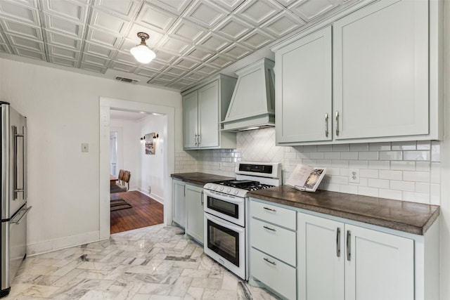 kitchen with custom exhaust hood, backsplash, stainless steel fridge, and range with two ovens