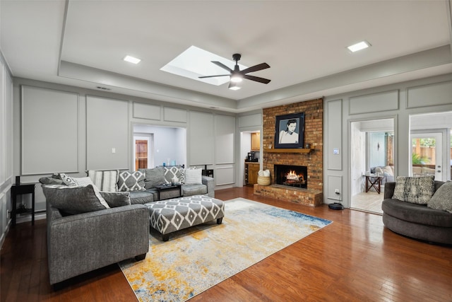 living room featuring a skylight, ceiling fan, a tray ceiling, dark hardwood / wood-style flooring, and a fireplace