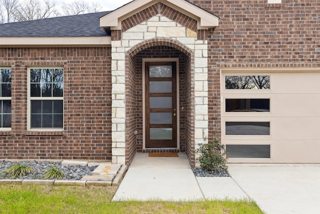 property entrance with brick siding, an attached garage, roof with shingles, and driveway