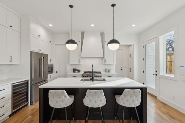 kitchen with custom exhaust hood, appliances with stainless steel finishes, an island with sink, and white cabinetry