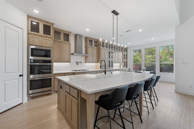 kitchen featuring stainless steel appliances, wall chimney range hood, a large island with sink, and light wood-style floors