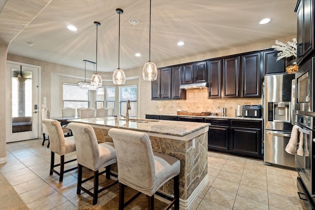 kitchen featuring pendant lighting, stainless steel appliances, an island with sink, sink, and light stone counters