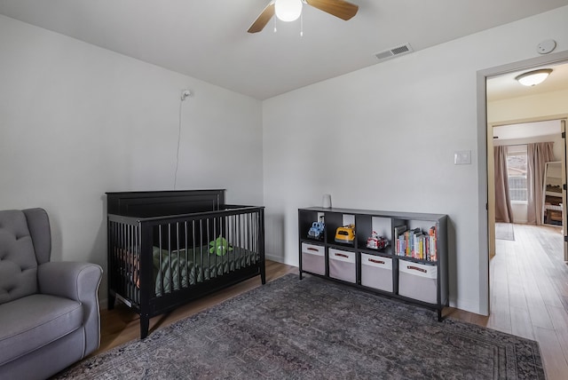 bedroom with ceiling fan, a nursery area, and hardwood / wood-style floors