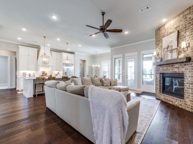 living room with crown molding, a brick fireplace, ceiling fan with notable chandelier, and dark wood-type flooring