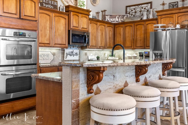 kitchen featuring light stone countertops, a kitchen island with sink, decorative backsplash, a kitchen breakfast bar, and stainless steel appliances