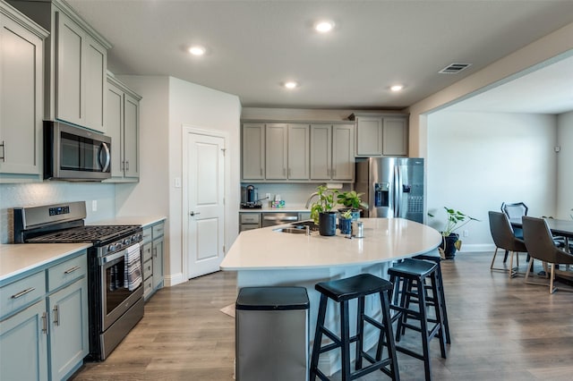 kitchen with appliances with stainless steel finishes, gray cabinetry, a kitchen bar, a kitchen island with sink, and light hardwood / wood-style floors