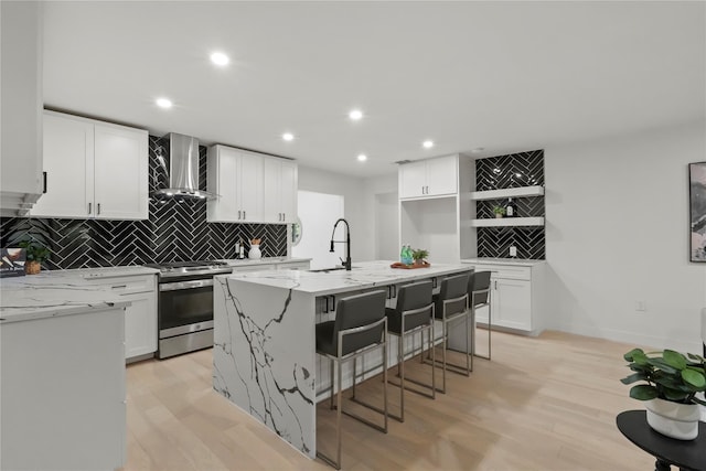 kitchen featuring wall chimney range hood, an island with sink, stainless steel range oven, and white cabinets