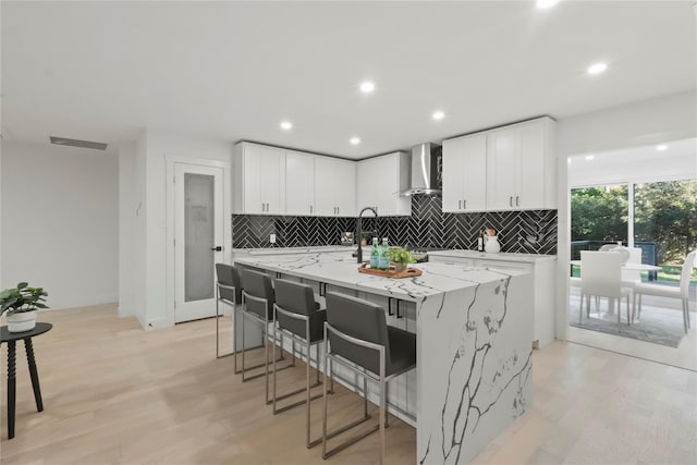 kitchen featuring white cabinets, light stone countertops, a breakfast bar, wall chimney range hood, and a large island with sink