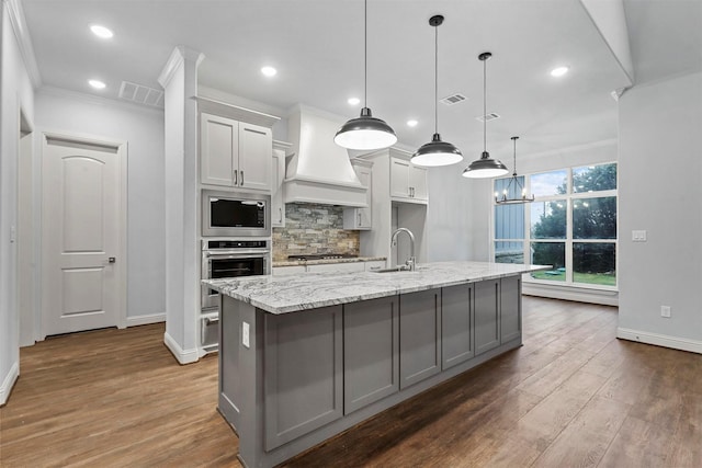 kitchen featuring appliances with stainless steel finishes, premium range hood, sink, decorative light fixtures, and a large island with sink