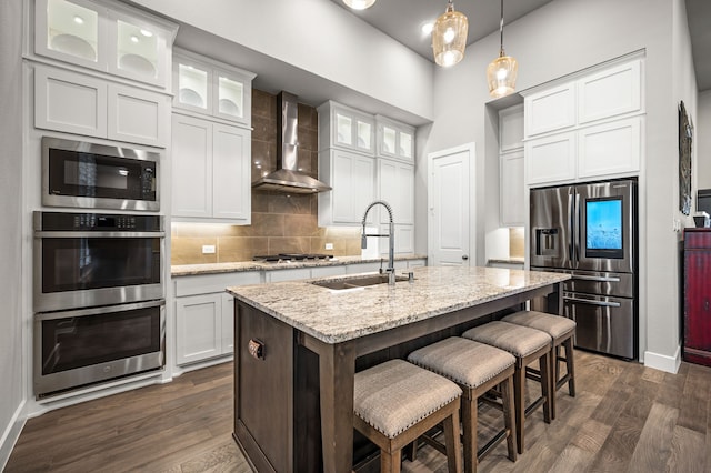 kitchen featuring light stone countertops, wall chimney range hood, stainless steel appliances, an island with sink, and sink